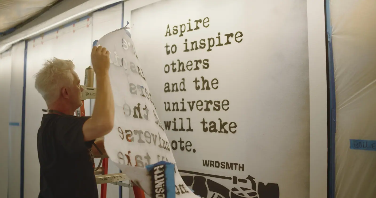 WRDSMTH on a ladder, pasting up “Aspire to inspire others and the universe will take note.”