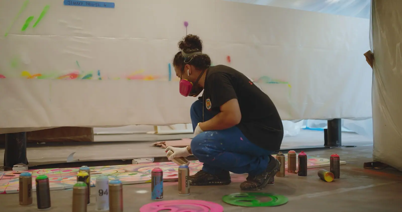 An artist in a respirator, crouched among spray cans, painting a canvas on the floor